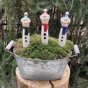 Könnte beinhalten: Drei Keramik-Schneemannfiguren mit grünen, roten und blauen Schals und Hüten, platziert in einer Metallwanne mit grünem Moos. Die Schneemänner haben schwarze Knopfaugen und Karottennasen. Die Wanne hat zwei Griffe.