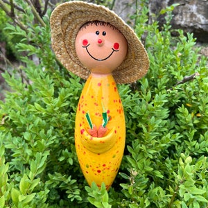 May include: A whimsical garden stake featuring a smiling figure with a straw hat. The stake is yellow with red and green polka dots, holding a pocket with two orange carrots. The face has rosy cheeks and a cheerful expression.