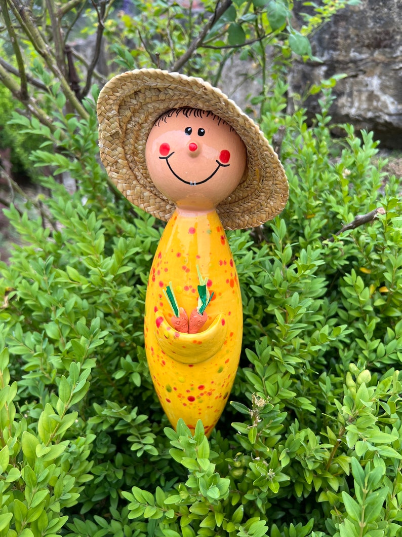 May include: A whimsical garden stake featuring a smiling figure with a straw hat. The stake is yellow with red and green polka dots, holding a pocket with two orange carrots. The face has rosy cheeks and a cheerful expression.