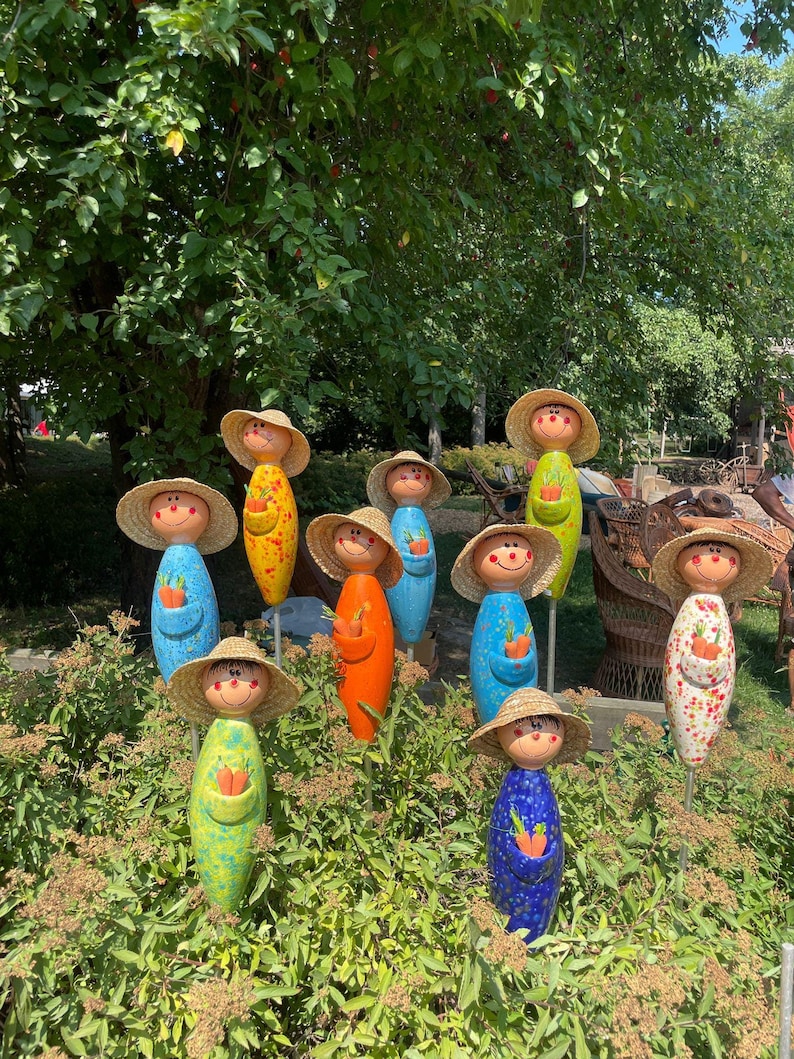 May include: A collection of colorful garden stakes shaped like smiling figures, each with a straw hat and holding a carrot. The stakes are painted in various colors, including blue, yellow, orange, and green, and are placed in a garden setting.