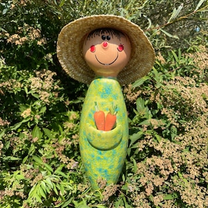 May include: A whimsical garden stake featuring a smiling face with a straw hat. The stake has a green and blue speckled body with a pocket holding two orange carrots. The garden stake is surrounded by green foliage.