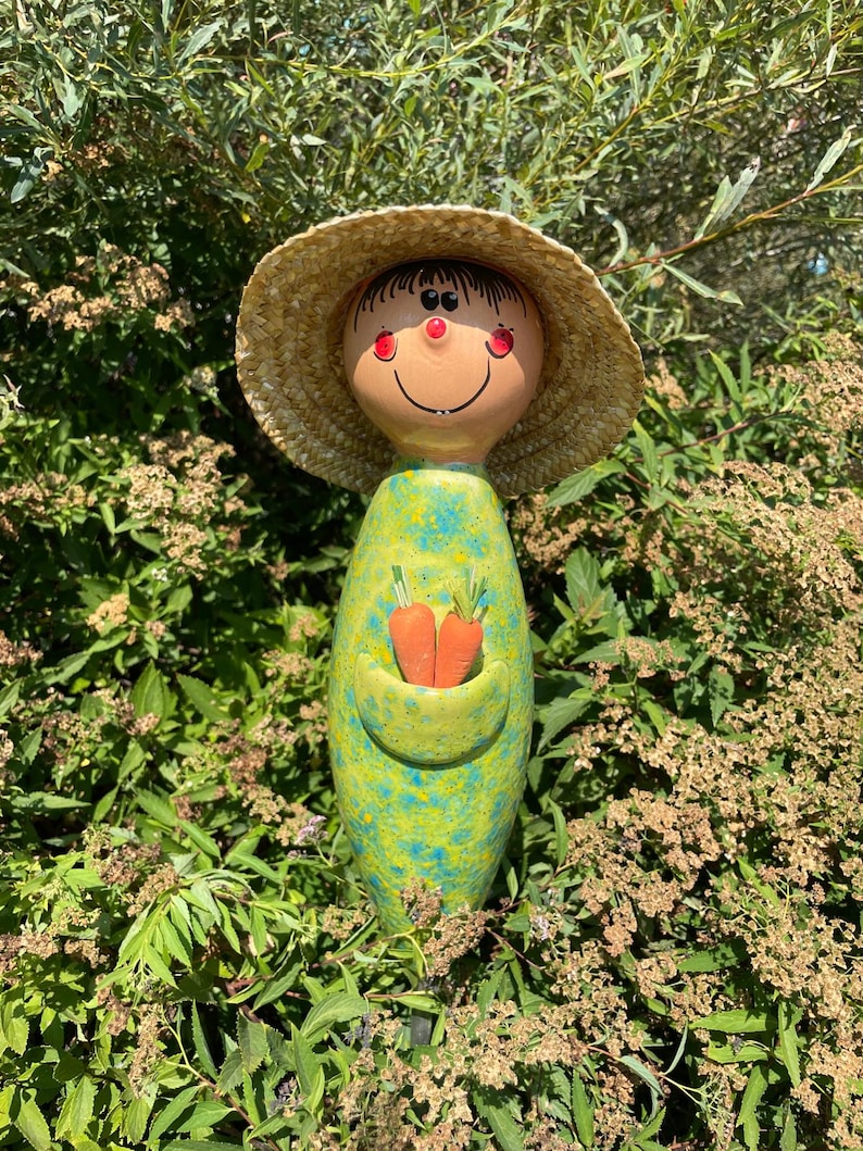 May include: A whimsical garden stake featuring a smiling face with a straw hat. The stake has a green and blue speckled body with a pocket holding two orange carrots. The garden stake is surrounded by green foliage.