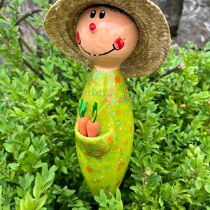 May include: A whimsical garden stake featuring a smiling figure with a straw hat and a green dress speckled with orange dots. The figure holds two orange carrot figurines in a pocket. The stake is set amidst green foliage.