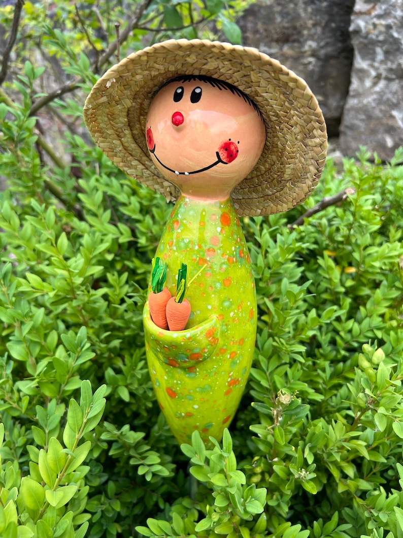 May include: A whimsical garden stake featuring a smiling figure with a straw hat and a green dress speckled with orange dots. The figure holds two orange carrot figurines in a pocket. The stake is set amidst green foliage.