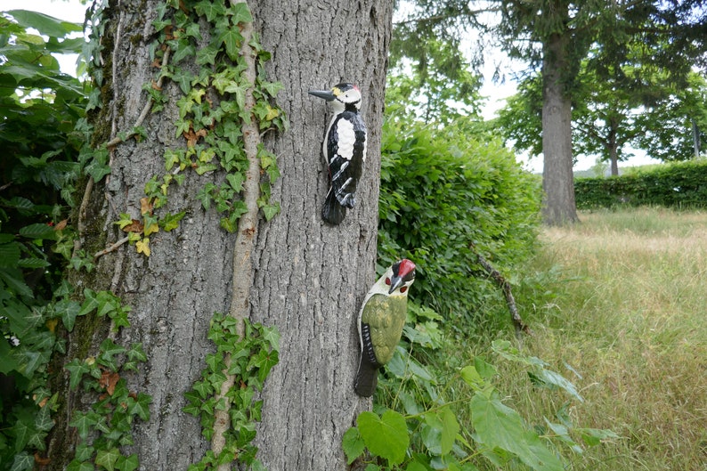 Peut inclure: Deux figurines d'oiseaux en bois fix&eacute;es &agrave; un tronc d'arbre. L'oiseau de gauche est noir et blanc avec un bec rouge. L'oiseau de droite est vert avec une t&ecirc;te rouge et un bec noir.