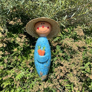 May include: A whimsical ceramic garden figurine of a smiling figure wearing a straw hat. The figure has a blue body with a pocket holding two orange carrots. The face is tan with red cheeks and a red smile.