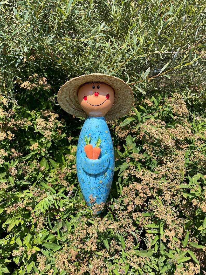 May include: A whimsical ceramic garden figurine of a smiling figure wearing a straw hat. The figure has a blue body with a pocket holding two orange carrots. The face is tan with red cheeks and a red smile.