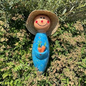 May include: A whimsical garden statue of a smiling figure with a straw hat. The blue ceramic body has orange speckles and holds two small orange carrots. The face is painted with a cheerful expression.