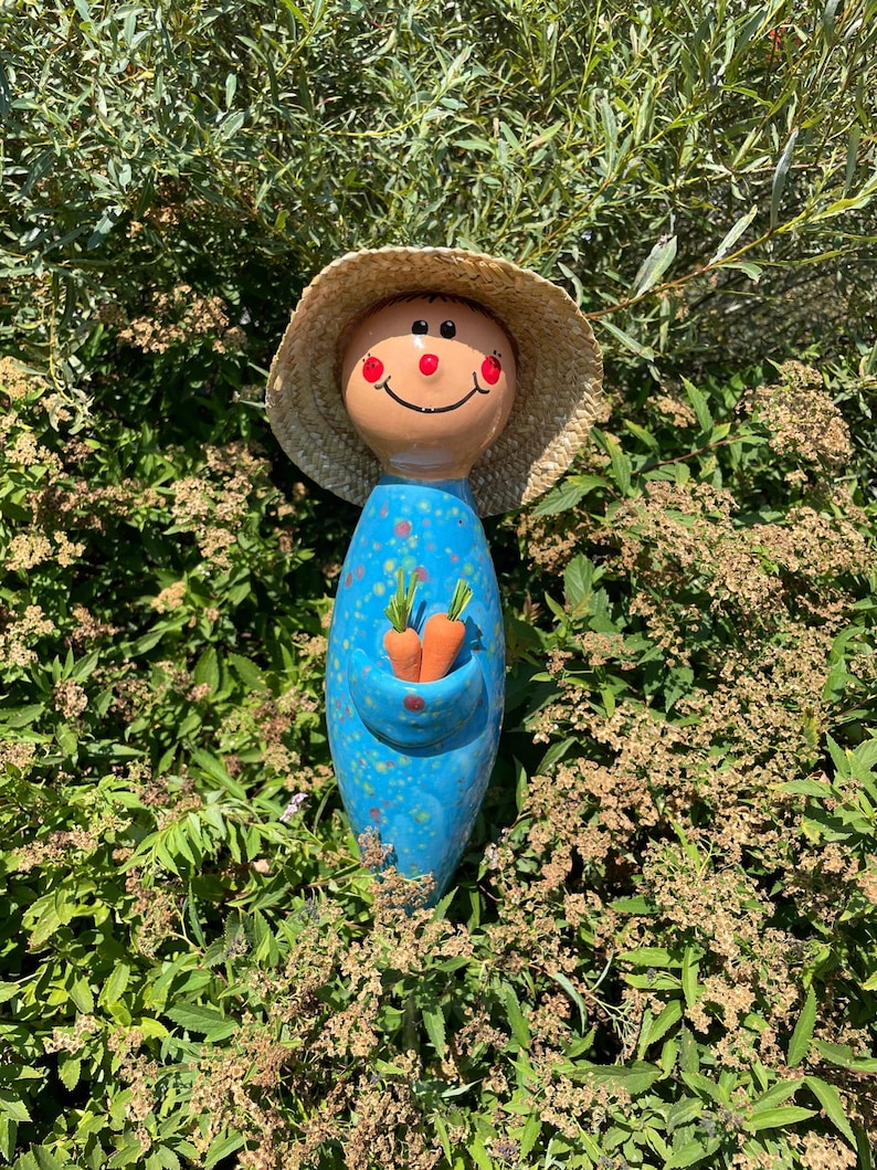 May include: A whimsical garden statue of a smiling figure with a straw hat. The blue ceramic body has orange speckles and holds two small orange carrots. The face is painted with a cheerful expression.