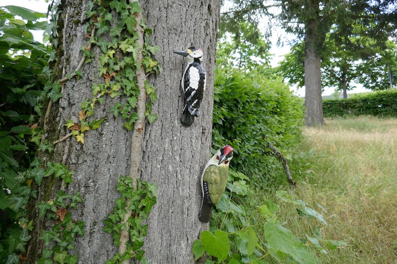 Peut inclure: Deux figurines d'oiseaux en bois fix&eacute;es &agrave; un tronc d'arbre. L'oiseau de gauche est noir et blanc avec un bec rouge. L'oiseau de droite est vert avec une t&ecirc;te rouge et des yeux noirs.