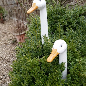 Könnte beinhalten: Zwei weiße Keramik-Gänsefiguren mit orangefarbenen Schnäbeln sind teilweise in einem grünen Busch versteckt. Die Gänse schauen in verschiedene Richtungen.