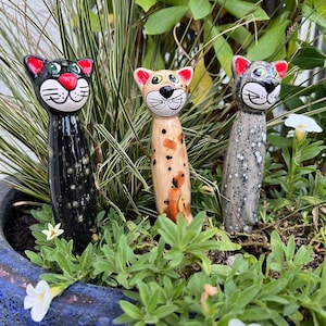 Könnte beinhalten: Drei Keramik-Katzenfiguren mit verschiedenen Mustern und Farben. Die Katzen stehen aufrecht mit nach oben geneigten Köpfen. Die Katze links ist schwarz mit weißen Flecken, die Katze in der Mitte ist braun mit orangefarbenen Flecken und die Katze rechts ist grau mit weißen Flecken.