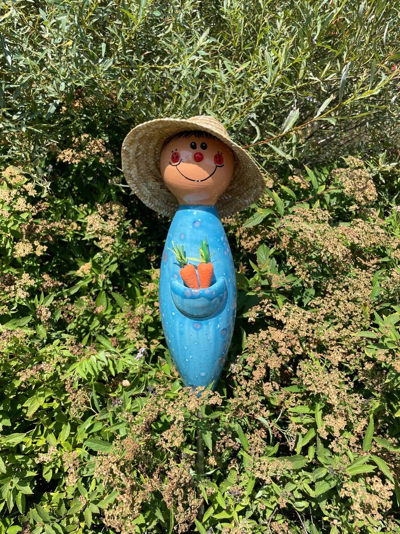 May include: A whimsical garden stake featuring a smiling face with a straw hat. The blue body has a pocket with two orange carrot decorations. The stake is surrounded by green foliage.