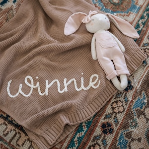 May include: A brown knitted baby blanket with the name "Winnie" embroidered in white thread. A pink bunny plush toy with overalls is lying on top of the blanket.