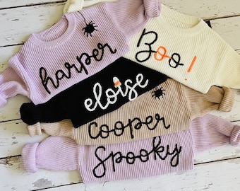 May include: A collection of knit sweaters in various colors, including lavender, cream, black, and tan. Each sweater features a name or word in a contrasting color, such as "Harper," "Boo," "Eloise," "Cooper," and "Spooky." Some sweaters have spider accents.
