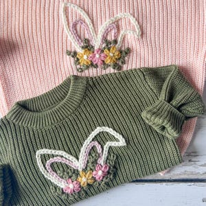 May include: Two knitted sweaters, one pink and one olive green, are displayed. Each sweater features a crocheted bunny ear design with floral accents. The bunny ears are white with pink and purple details, and the flowers are yellow and pink.