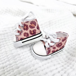 May include: A pair of miniature canvas sneakers with a pink leopard print pattern. The shoes have white laces, a white toe cap, and a black stripe along the sole. The sneakers are designed for a small child or doll.