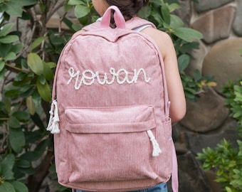 May include: A pink corduroy backpack with white rope detailing and the name "Rowan" embroidered on the front.