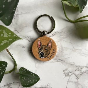 May include: A wooden keychain with a painted portrait of a brown and white dog. The dog's name, "KAISER", is written on the keychain.