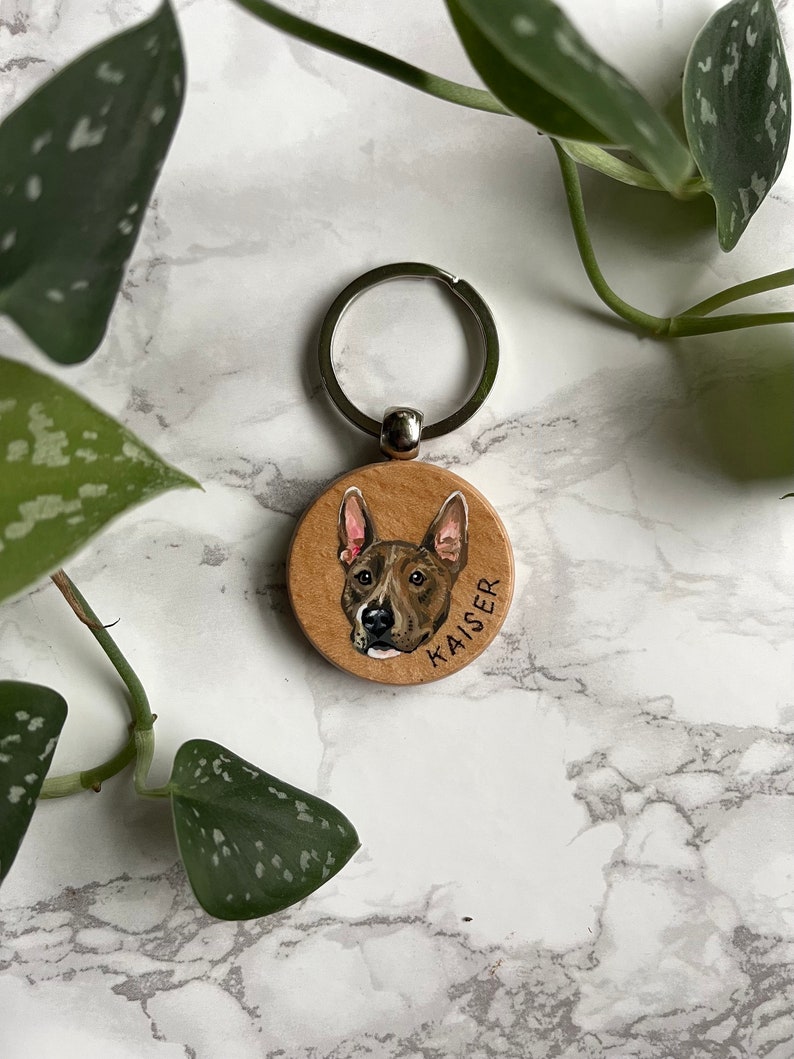 May include: A wooden keychain with a painted portrait of a brown and white dog. The dog's name, "KAISER", is written on the keychain.