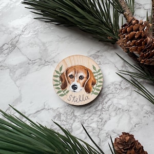 May include: A round wooden ornament featuring a painted beagle dog portrait with the name "Stella" in cursive. The dog is brown and white, surrounded by green leaves. The background includes pine needles and a marble surface.