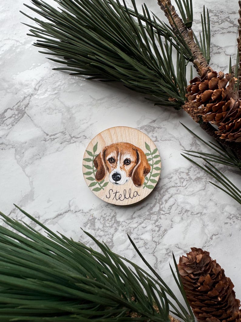 May include: A round wooden ornament featuring a painted beagle dog portrait with the name "Stella" in cursive. The dog is brown and white, surrounded by green leaves. The background includes pine needles and a marble surface.