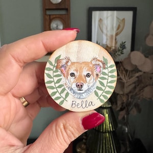 May include: A round wooden ornament featuring a detailed portrait of a dog with brown and white fur, surrounded by green leaves. The dog's name, "Bella," is written in black script below the portrait. The ornament is held in a hand.