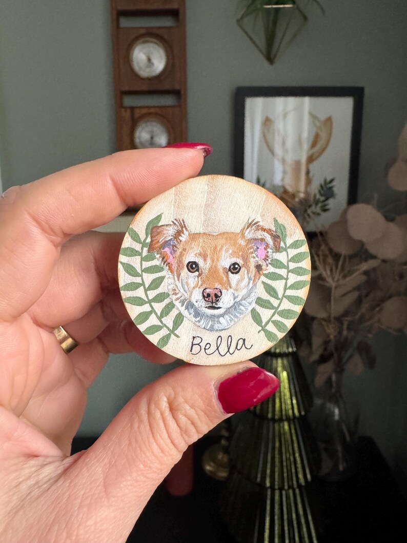 May include: A round wooden ornament featuring a detailed portrait of a dog with brown and white fur, surrounded by green leaves. The dog's name, "Bella," is written in black script below the portrait. The ornament is held in a hand.