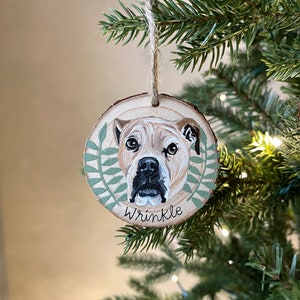 Puede incluir: Un adorno redondo de madera con un retrato pintado de un perro. El perro tiene pelo marr&oacute;n y blanco y una nariz negra. El adorno est&aacute; decorado con hojas verdes y el nombre "Wrinkle" est&aacute; escrito debajo del retrato del perro.