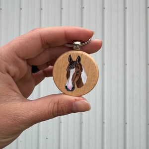 May include: A wooden keychain with a painted image of a brown and white horse's head. The horse has a pink nose and a black mane.