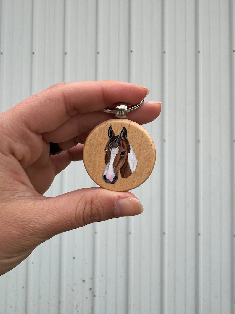 May include: A wooden keychain with a painted image of a brown and white horse's head. The horse has a pink nose and a black mane.