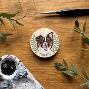 May include: A round wooden coaster featuring a painted portrait of a dog with brown, white, and black fur, surrounded by green leaves. The word "Toasty" is written below the dog's portrait. A paintbrush and paint palette are also visible.
