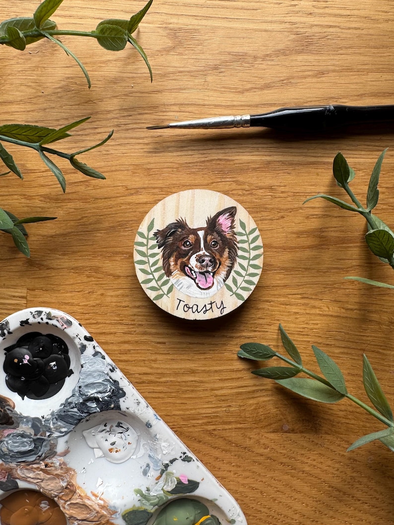 May include: A round wooden coaster featuring a painted portrait of a dog with brown, white, and black fur, surrounded by green leaves. The word "Toasty" is written below the dog's portrait. A paintbrush and paint palette are also visible.