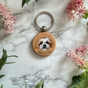 May include: A wooden keychain with a painted portrait of a dog. The dog has white fur with brown markings and is looking directly at the camera. The keychain is round and has the name "Wolfie" written on it.