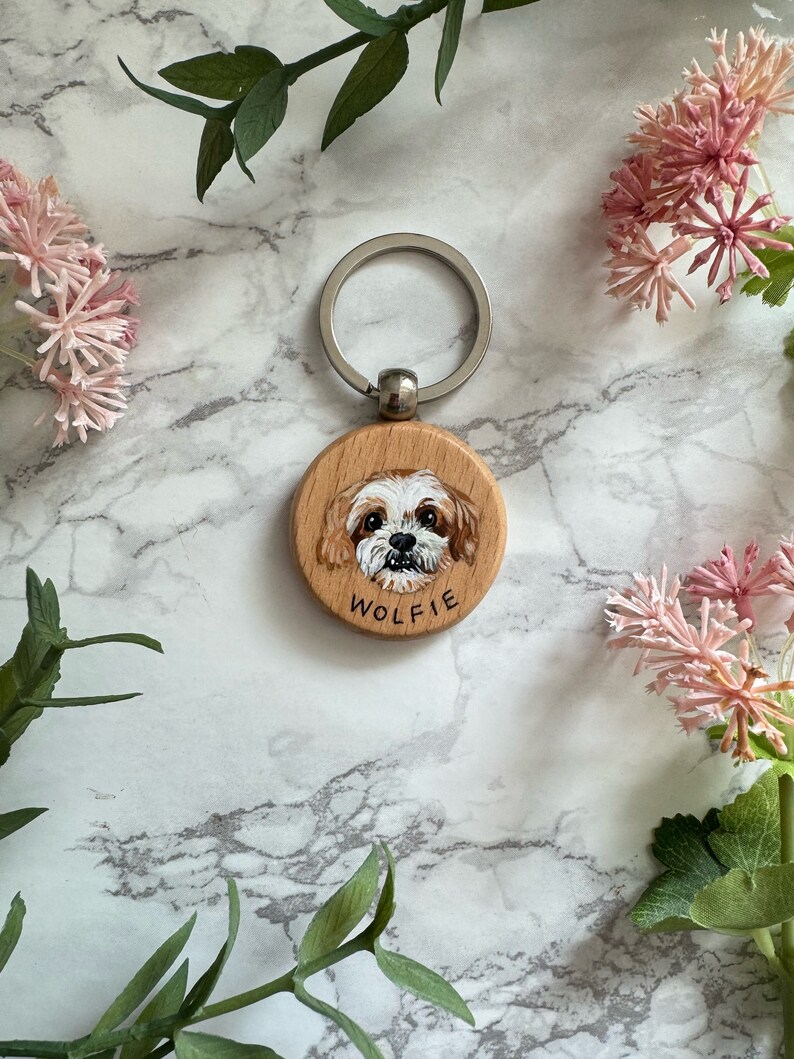 May include: A wooden keychain with a painted portrait of a dog. The dog has white fur with brown markings and is looking directly at the camera. The keychain is round and has the name "Wolfie" written on it.