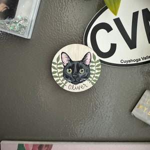 May include: A round wooden magnet featuring a black cat portrait with green eyes, surrounded by a green leaf design. The cat's name, "Oliver," is written below. The magnet is affixed to a gray surface.