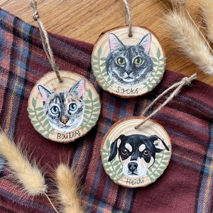 Puede incluir: Tres adornos de madera con retratos pintados de mascotas. Los adornos son redondos y tienen una veta de madera r&uacute;stica. El primer adorno presenta un gato atigrado gris con ojos verdes y el nombre "Socks" escrito debajo. El segundo adorno presenta un gato atigrado gris y blanco con ojos azules y el nombre "Bailey" escrito debajo. El tercer adorno presenta un perro negro y marr&oacute;n con ojos marrones y el nombre "Heidi" escrito debajo.