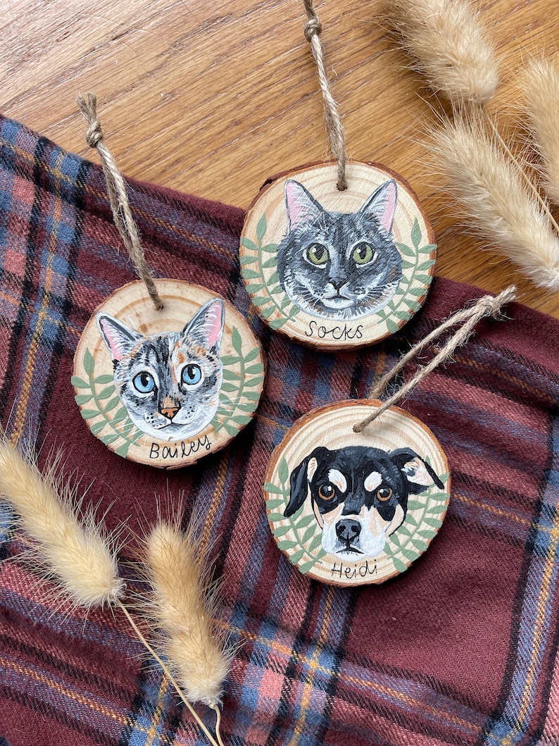 Puede incluir: Tres adornos de madera con retratos pintados de mascotas. Los adornos son redondos y tienen una veta de madera r&uacute;stica. El primer adorno presenta un gato atigrado gris con ojos verdes y el nombre "Socks" escrito debajo. El segundo adorno presenta un gato atigrado gris y blanco con ojos azules y el nombre "Bailey" escrito debajo. El tercer adorno presenta un perro negro y marr&oacute;n con ojos marrones y el nombre "Heidi" escrito debajo.