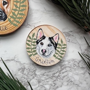 May include: Hand-painted wooden ornament featuring a dog's portrait, surrounded by green leaves. The dog has one blue eye and one brown eye. The name "Griffin" is written below the portrait. The ornament is made of wood and has a natural wood grain.