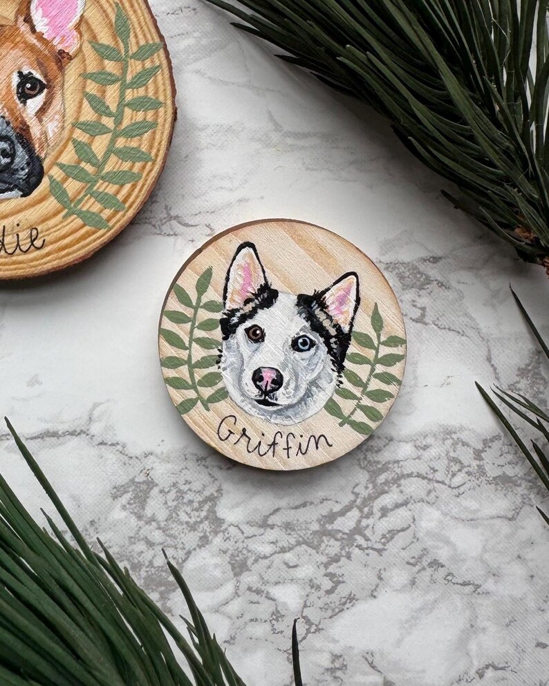 May include: Hand-painted wooden ornament featuring a dog's portrait, surrounded by green leaves. The dog has one blue eye and one brown eye. The name "Griffin" is written below the portrait. The ornament is made of wood and has a natural wood grain.