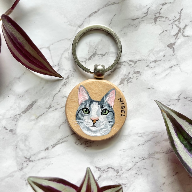 May include: A round wooden keychain with a painted portrait of a gray and white cat. The cat's name, "Nigel", is written on the keychain.
