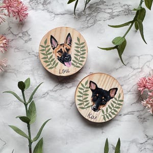 May include: Two round wooden pet portraits. One features a German Shepherd with the name "Lou" and the other a black and brown dog named "Kai". Each portrait is surrounded by a green leaf design.