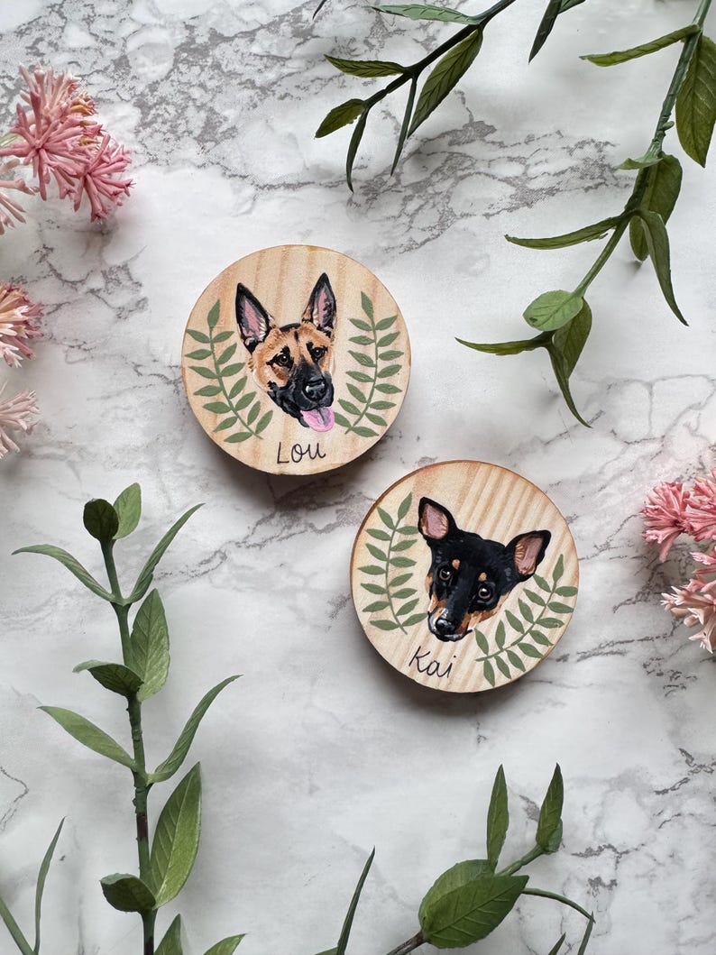 May include: Two round wooden pet portraits. One features a German Shepherd with the name "Lou" and the other a black and brown dog named "Kai". Each portrait is surrounded by a green leaf design.