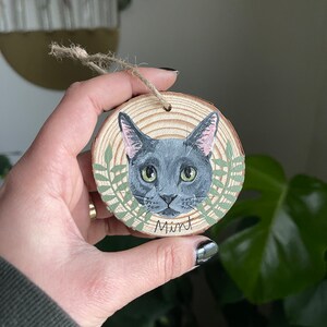 Puede incluir: Un adorno redondo de madera con un retrato pintado de un gato gris con ojos verdes. El gato est&aacute; rodeado de hojas verdes y el nombre "Mint" est&aacute; escrito debajo del gato.