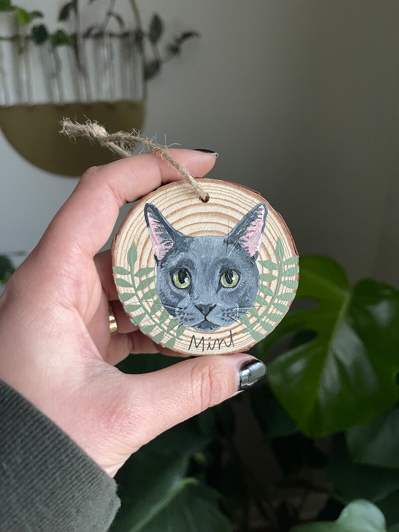 Puede incluir: Un adorno redondo de madera con un retrato pintado de un gato gris con ojos verdes. El gato est&aacute; rodeado de hojas verdes y el nombre "Mint" est&aacute; escrito debajo del gato.