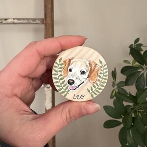 May include: A round wooden ornament featuring a painted portrait of a dog with tan and white fur. The dog's name, "Leo," is written below the portrait. Green leaves surround the dog portrait. The ornament is held in a person's hand.