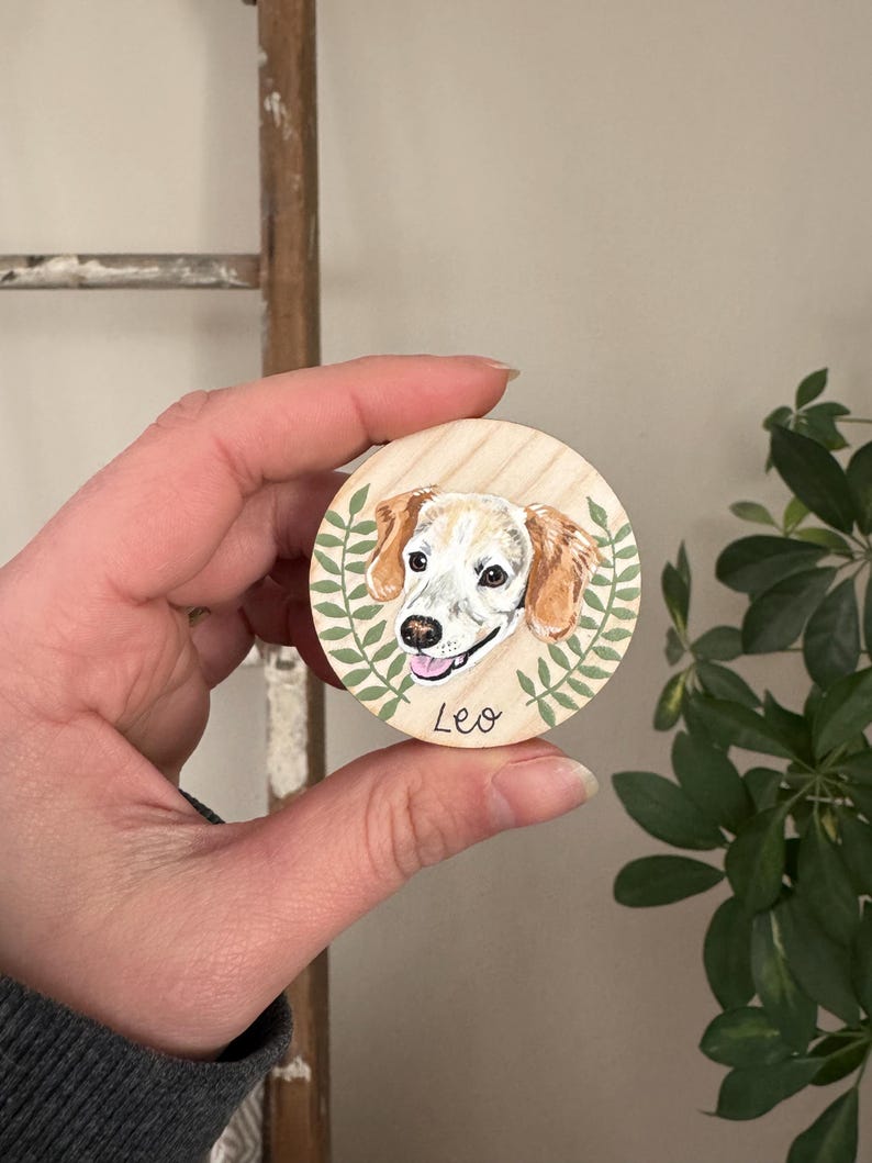 May include: A round wooden ornament featuring a painted portrait of a dog with tan and white fur. The dog's name, "Leo," is written below the portrait. Green leaves surround the dog portrait. The ornament is held in a person's hand.