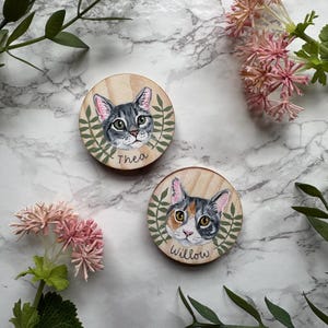 May include: Two wooden cat portraits. One features a gray tabby cat named Thea, the other a calico cat named Willow. Both are surrounded by a green leaf design. The background is a marble pattern with pink flowers and green leaves.