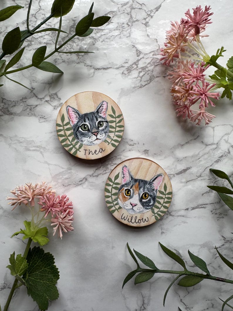 May include: Two wooden cat portraits. One features a gray tabby cat named Thea, the other a calico cat named Willow. Both are surrounded by a green leaf design. The background is a marble pattern with pink flowers and green leaves.