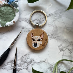 May include: A wooden keychain with a painted portrait of a dog. The dog is a brown and white Australian Cattle Dog with a black nose. The keychain is round and has a silver ring attached. The dog's name, "RODEO", is written in white on the keychain.